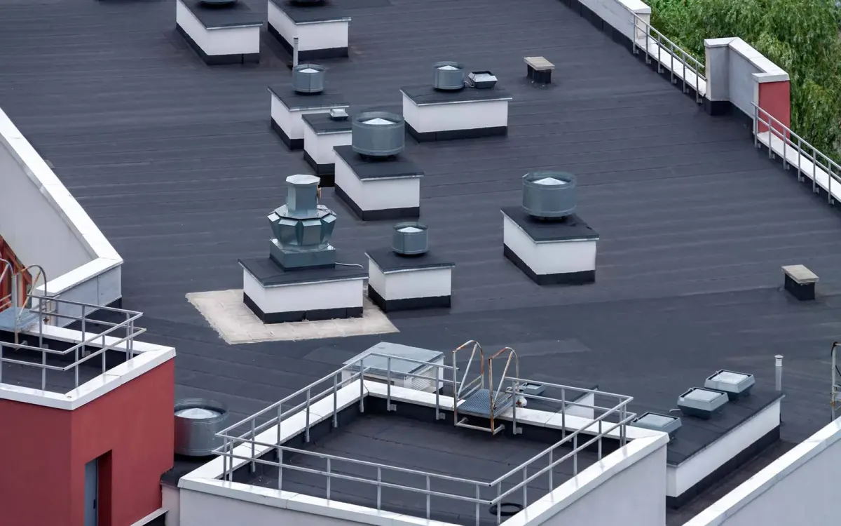 Aerial view of a dark bituminous low-slope roof with white curbs, round exhaust fans and rooftop guardrails