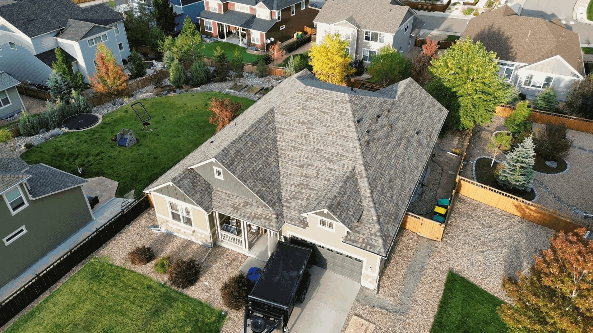 Aerial view of a single-story home's architectural shingle roof, with a dump trailer in the driveway and autumn trees in the surrounding yards.