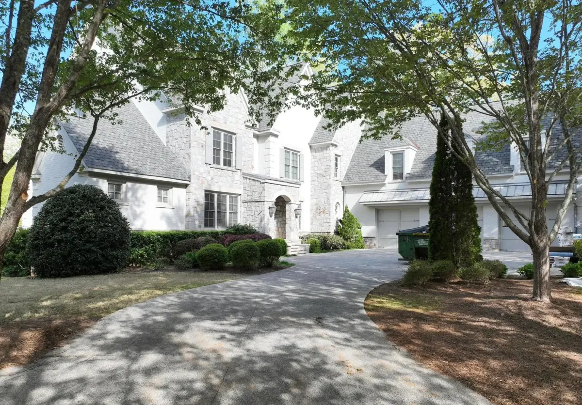 Large stone-front home with a gray shingle roof, seen up a tree-shaded driveway with a work dumpster by the garage