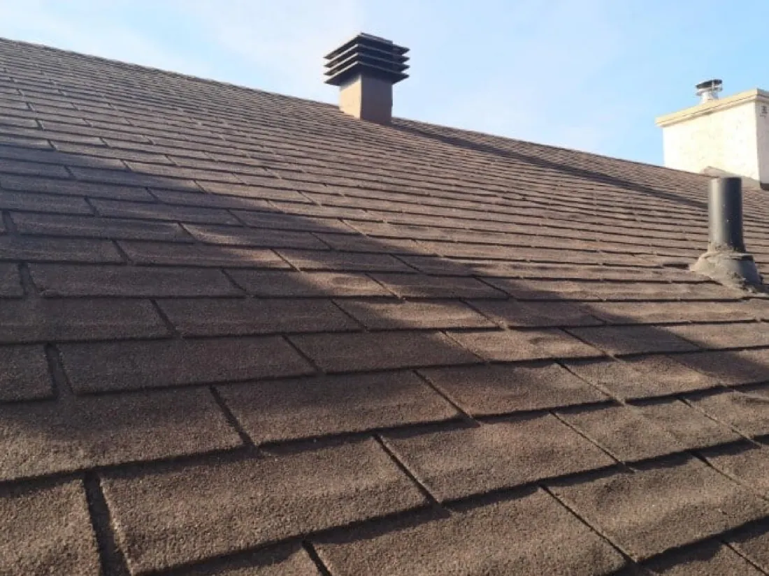 Looking up a slope of brown three-tab shingles toward a metal vent cap and a stucco chimney